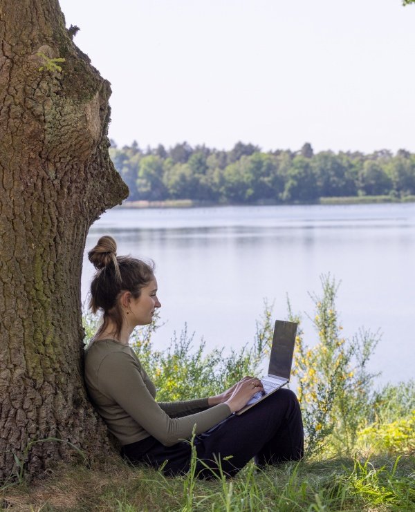 Femeie care lucrează la laptop rezemată de un copac în mijlocul naturii